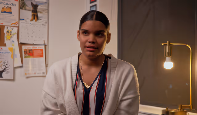 Woman with dark hair pulled back, wearing a white cardigan over a striped shirt, sitting indoors near a bulletin board and a lit lamp.