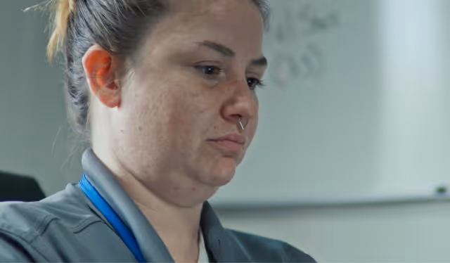 Close-up of a woman with a septum piercing and hair tied back, looking down with a neutral expression.
