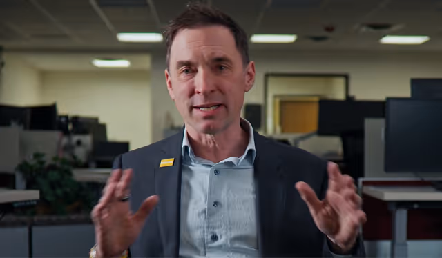 Man in a suit jacket speaking with hands raised in an office setting with computer monitors in the background.