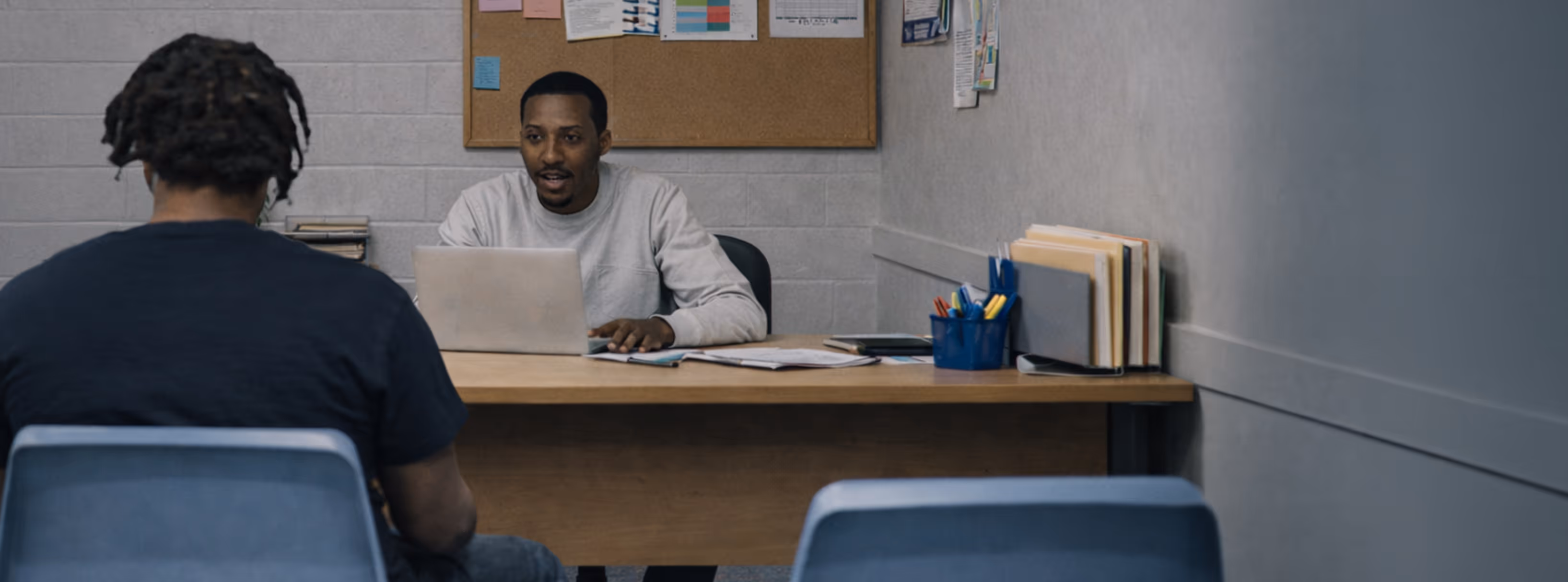 Two men are seated across from each other at a desk in a small office, one using a laptop while talking.