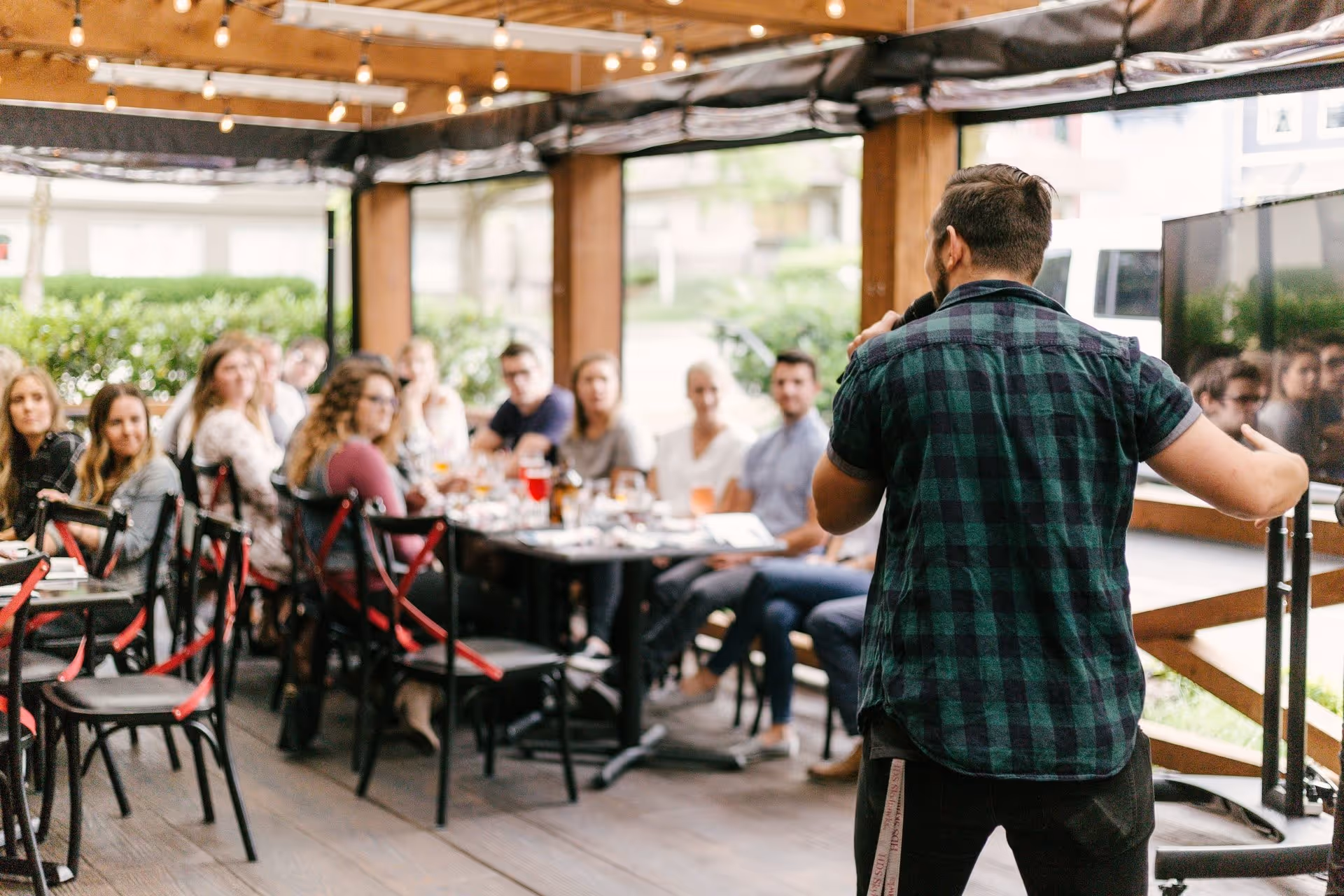 A person in a green plaid shirt speaks to a seated audience on an outdoor patio. The setting is casual and lively, with string lights overhead.