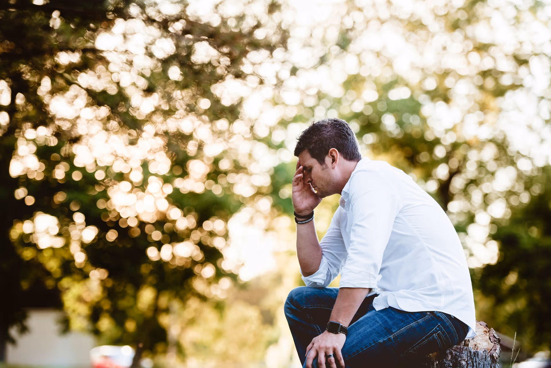 Man in white shirt and jeans sits on a tree stump in a park, covering his face with one hand, appearing stressed. Sunlight filters through leafy trees.