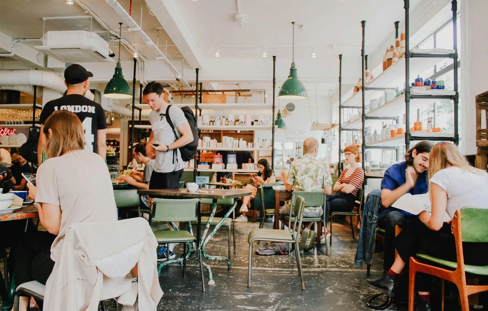 A busy cafe with people sitting at tables, chatting and using phones. Bright lighting, green pendant lamps, and shelves with colorful items create a lively atmosphere.