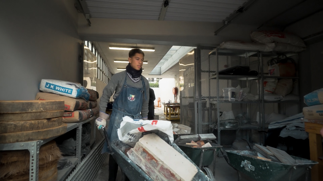 Man in plaid shirt speaks to an attentive audience at a casual, outdoor gathering with wooden decor and string lights, creating a warm atmosphere.