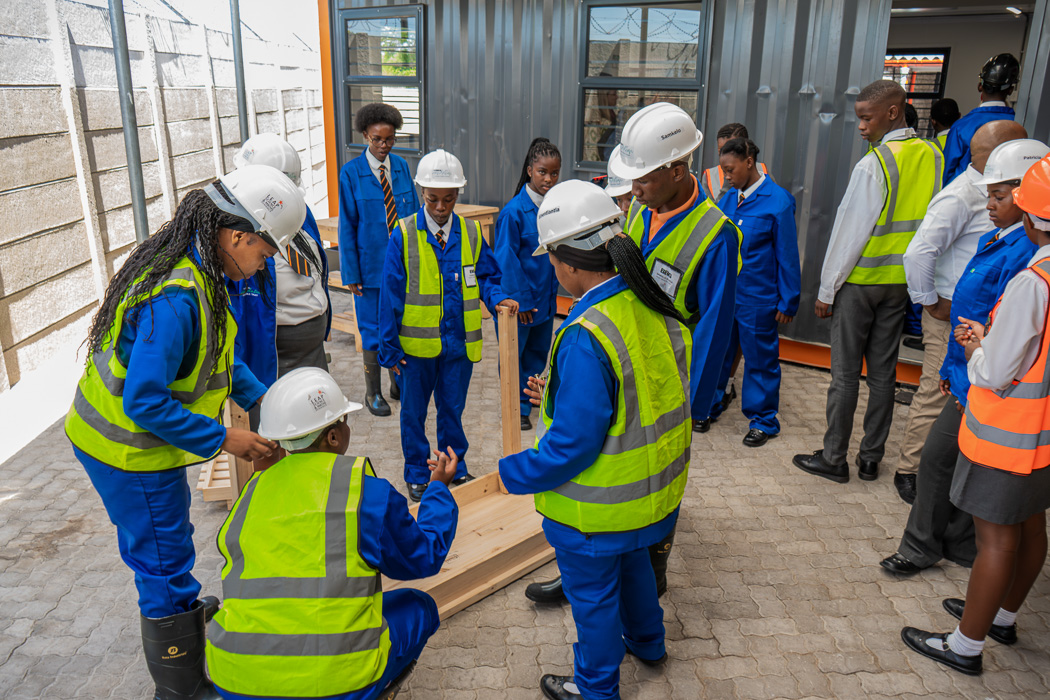 Two people tend to a vertical garden of black triangular pots with various green plants against a green and white grid, evoking a sense of urban gardening.