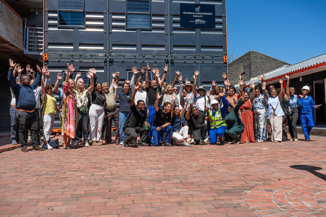Man in plaid shirt speaking to a seated audience in a cozy, well-lit room with string lights. The atmosphere is engaging and focused.