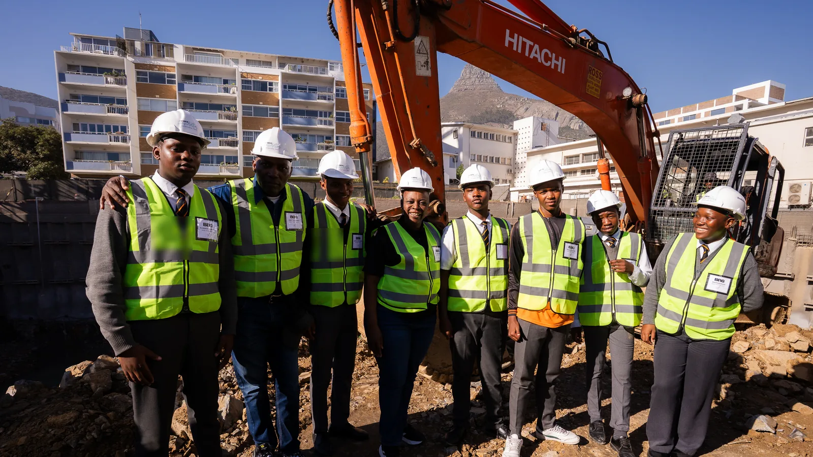 Hands On learners in hard hats and high-visibility vests standing with their teacher at a Cape Town construction site, Lion's Head in the background