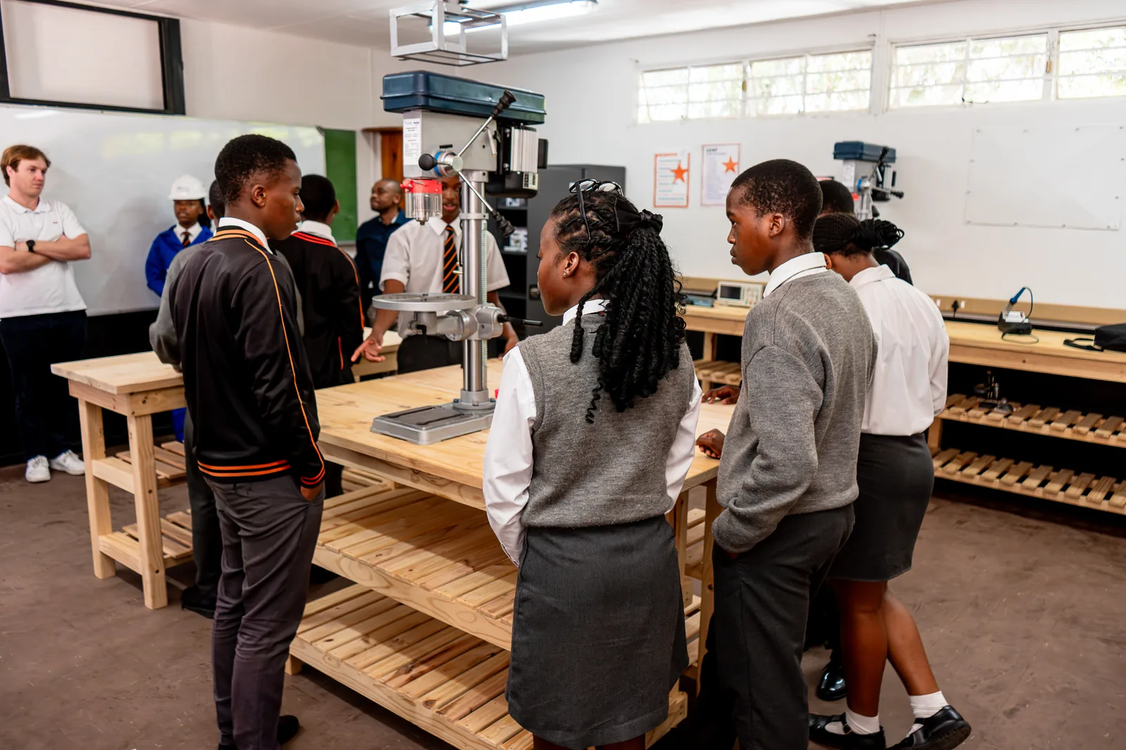 Learners gathered around a drill press on a wooden workbench inside a Hands On technical faculty workshop