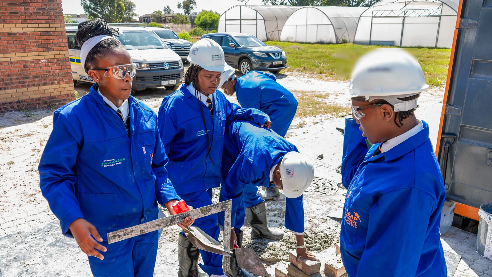Learners in blue overalls, hard hats, and safety glasses laying bricks with a builder's square and trowel during a practical lesson
