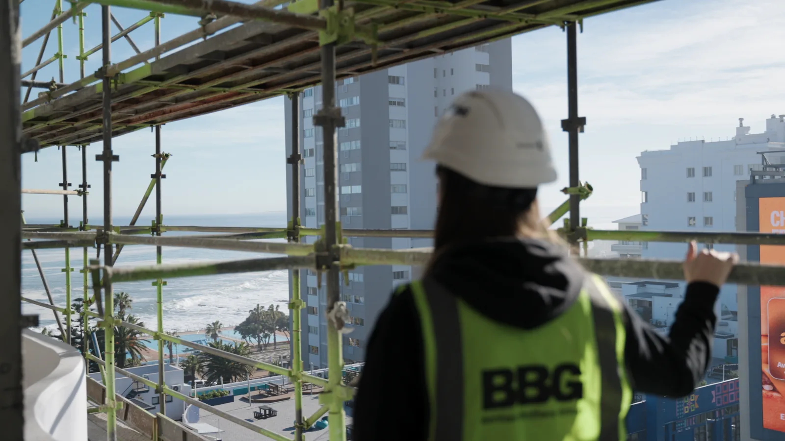 Marika Abrahams seen from behind in a hard hat on scaffolding, looking out over Sea Point and the Atlantic