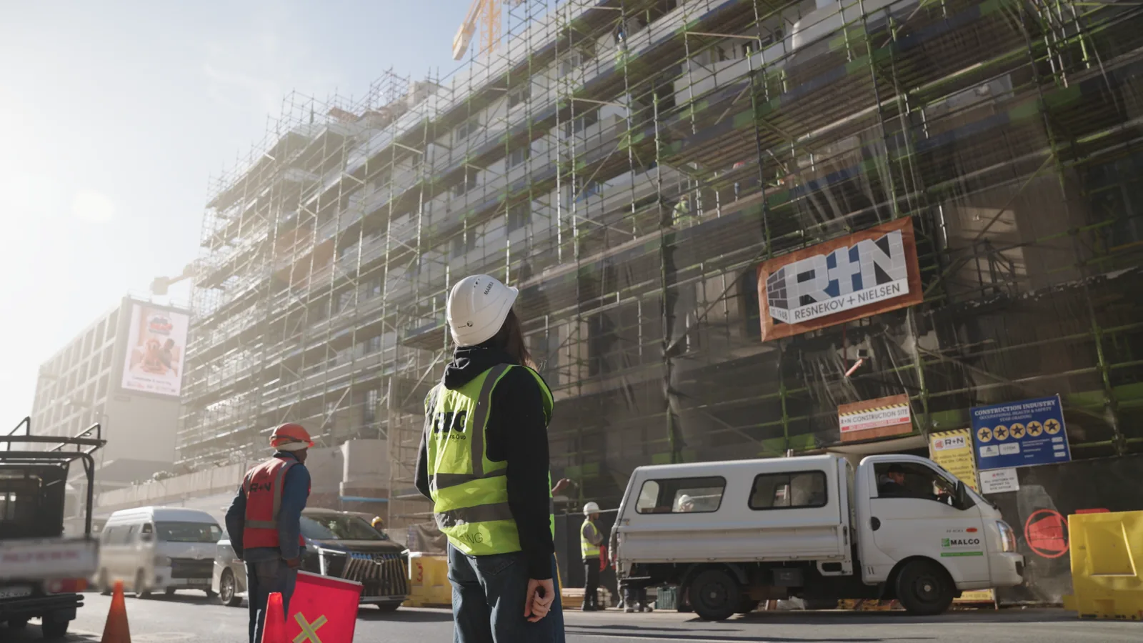 Marika Abrahams in a hard hat and high-visibility vest looking up at a scaffolded building on a working construction site