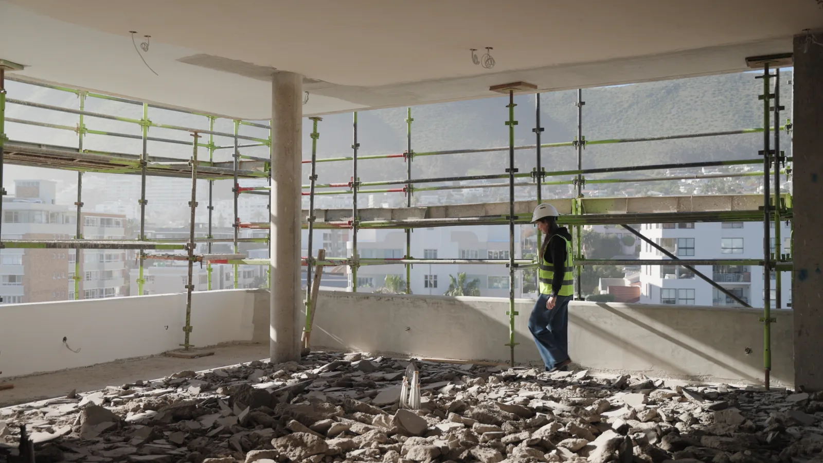 Marika Abrahams in a hard hat and high-visibility vest walking a scaffolded floor edge with Cape Town buildings behind