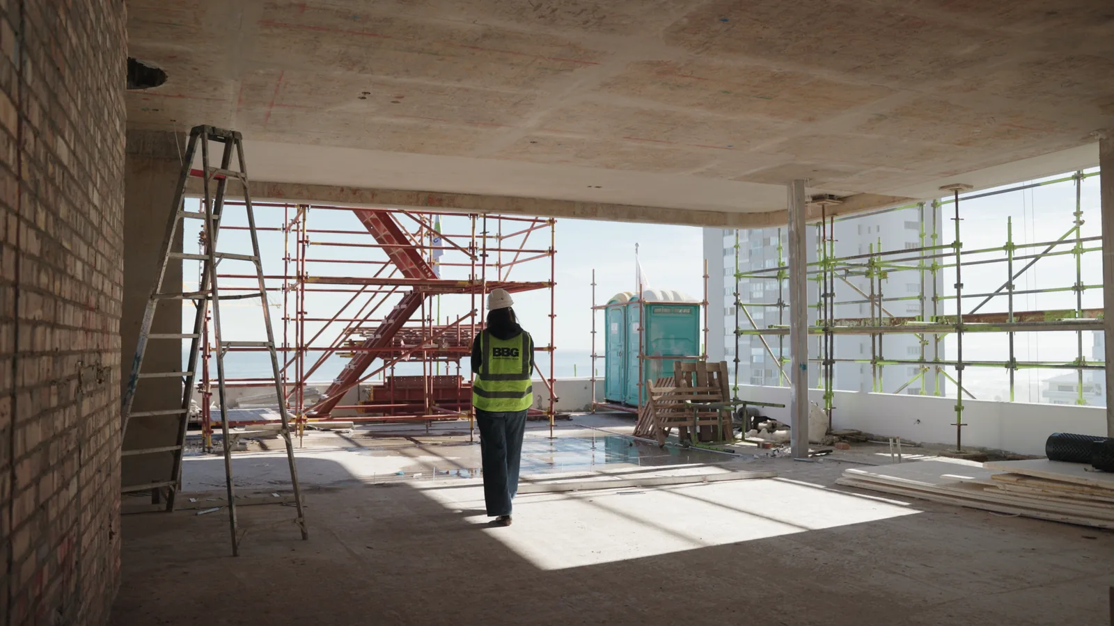 Marika Abrahams walking through an unfinished floor of a building under construction, scaffolding and sea views beyond