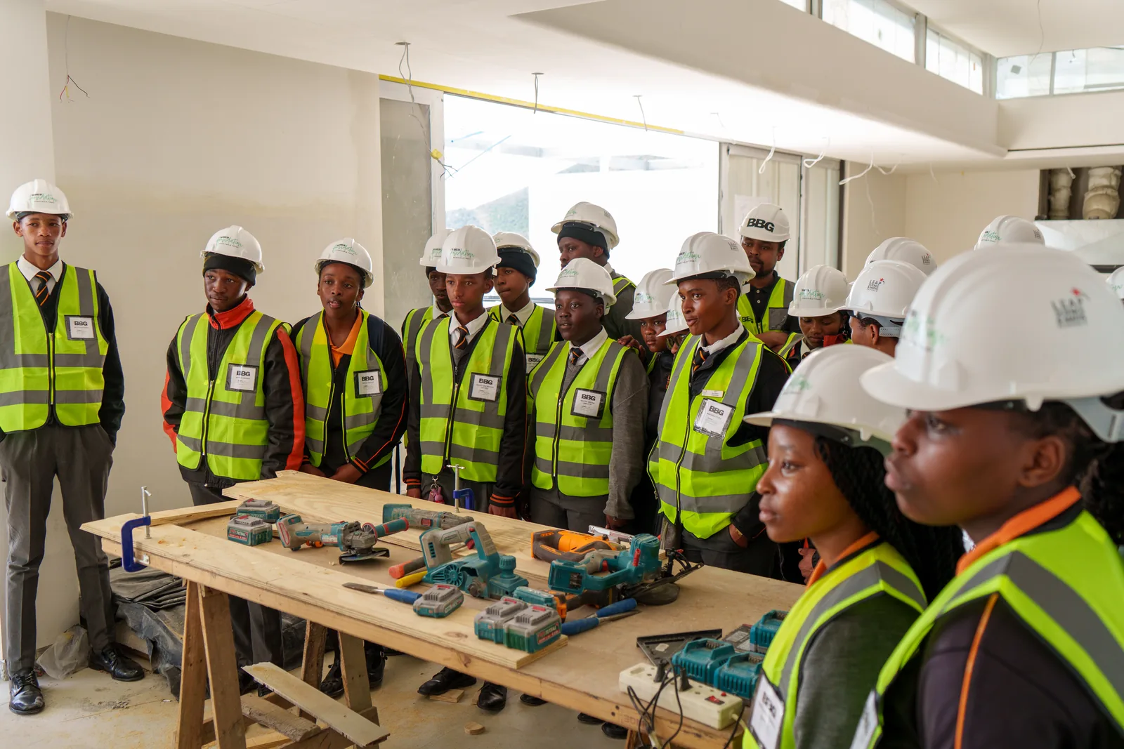 Learners in hard hats and high-visibility vests standing around a workbench of power tools during a construction site briefing
