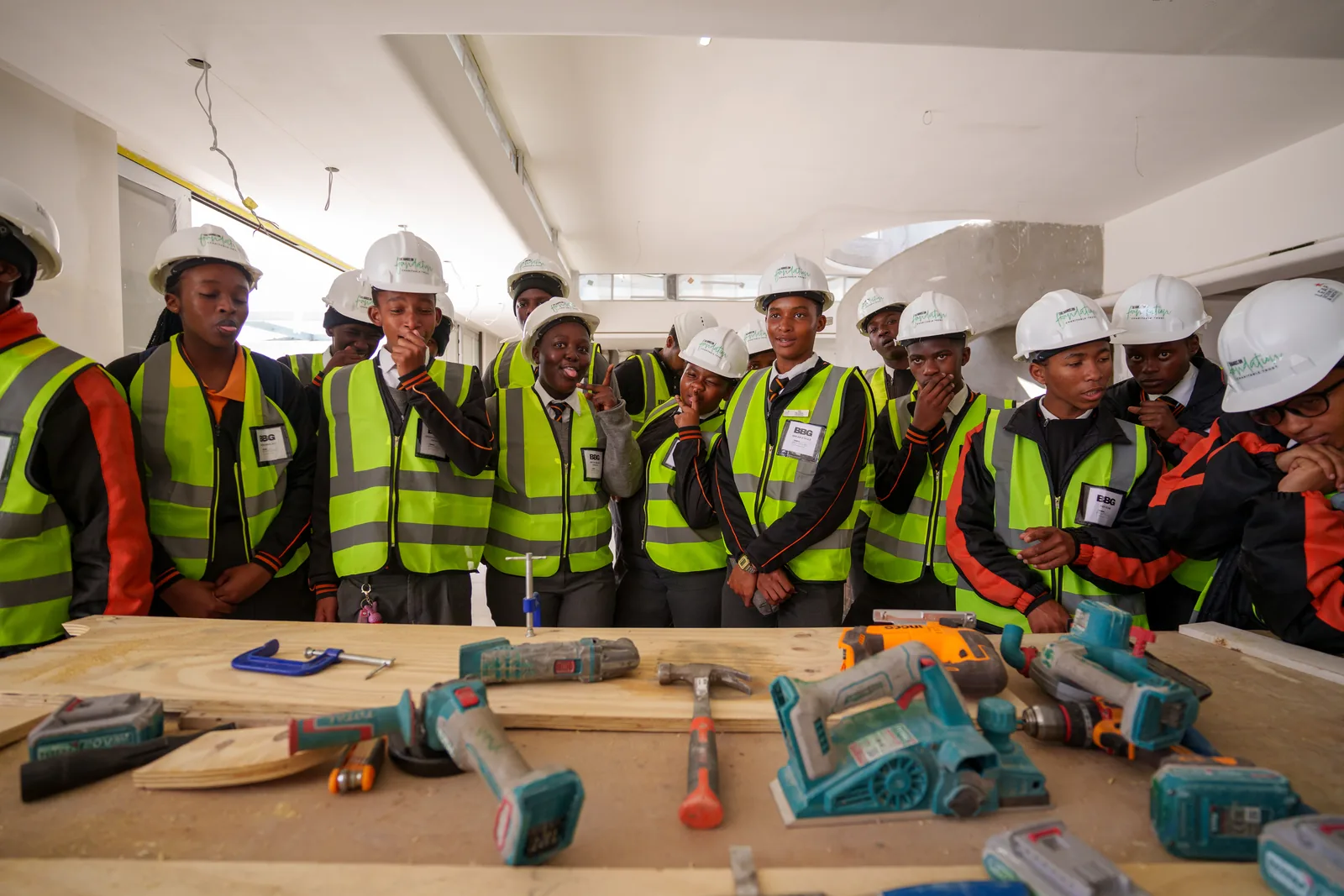Learners in hard hats and high-visibility vests gathered at a workbench of power tools on a working construction site