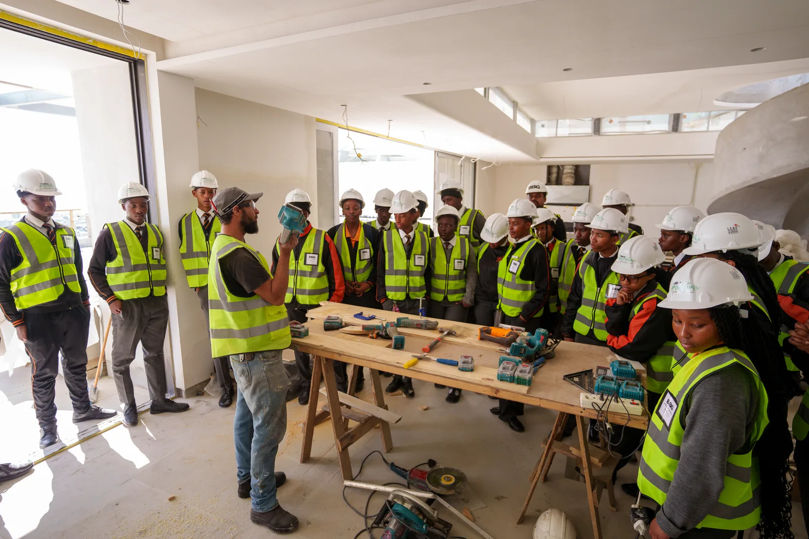 An artisan holding up a power tool while explaining it to a group of learners in hard hats on a live construction site