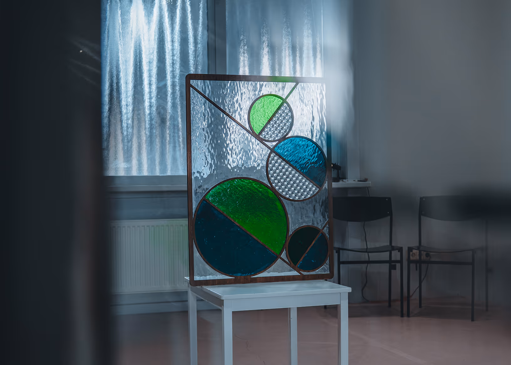 Stained glass panel with multiple green and blue circles on a white stool in a room with chairs and textured windows.