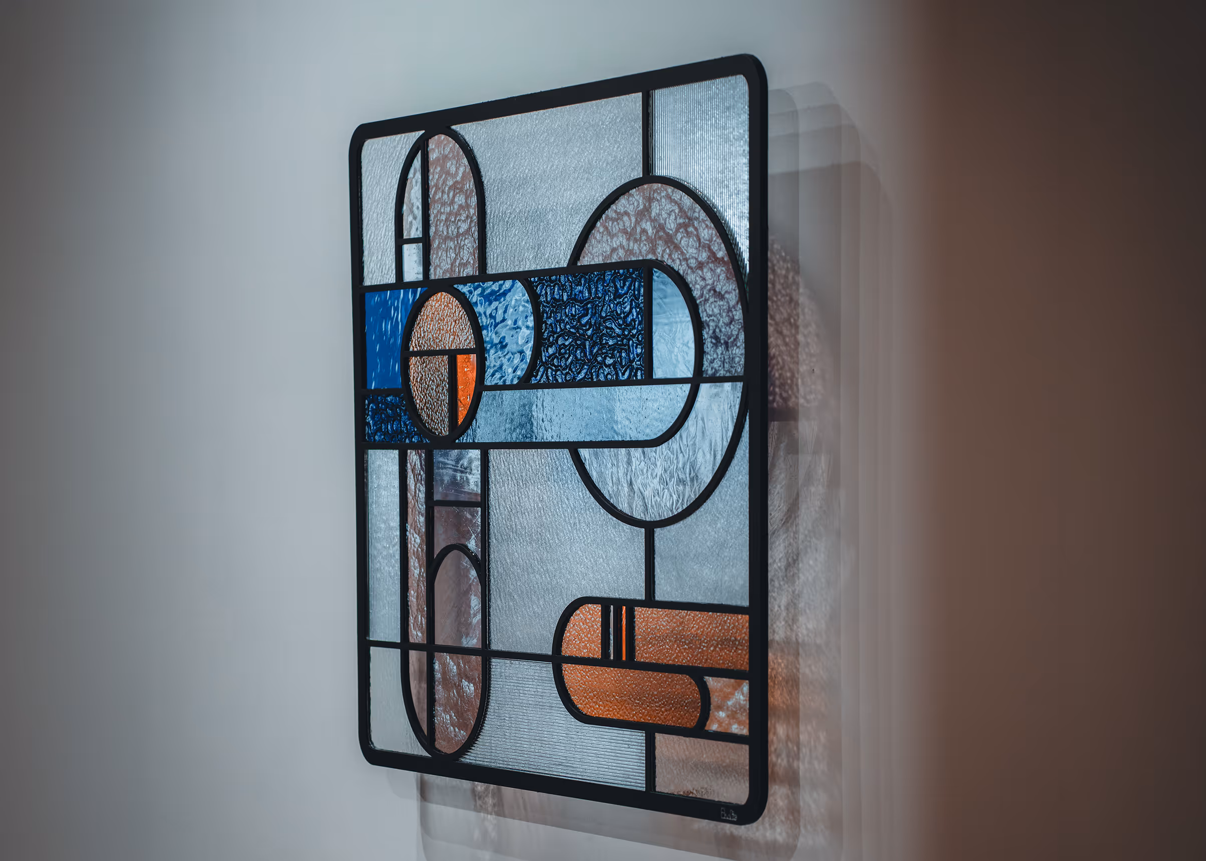 Rectangular stained glass panel with geometric shapes in blue, orange, and clear textured glass framed in black.