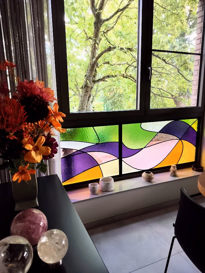 Room corner with a window featuring colorful stained glass panels in green, purple, yellow, pink, and white, looking out to green trees; a flower vase with orange and red flowers and crystal spheres are placed on a black table near the window.