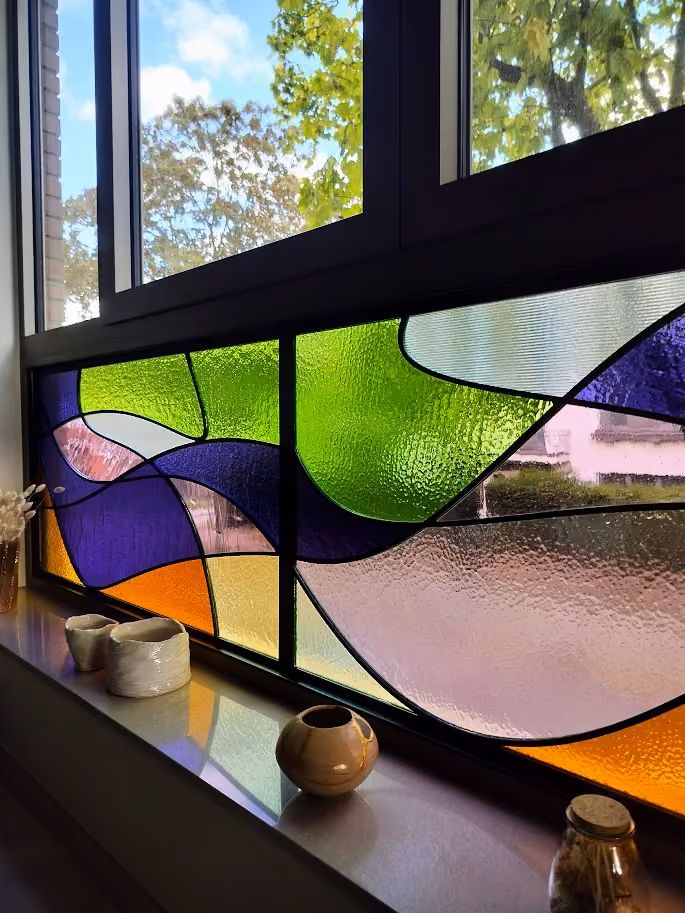 A window with colorful stained glass panels in shades of green, purple, orange, and textured translucent glass, with small ceramic pots placed on the window sill.