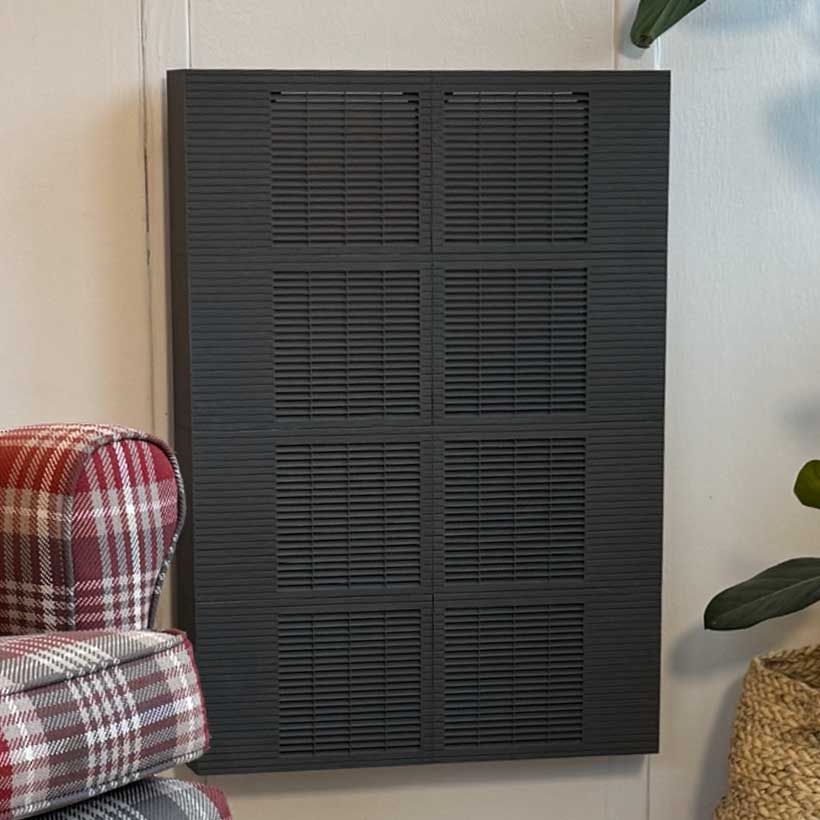 Black wall-mounted Trimley fan convector panel with horizontal slats, positioned between a red plaid armchair and a woven basket with green leaves nearby.