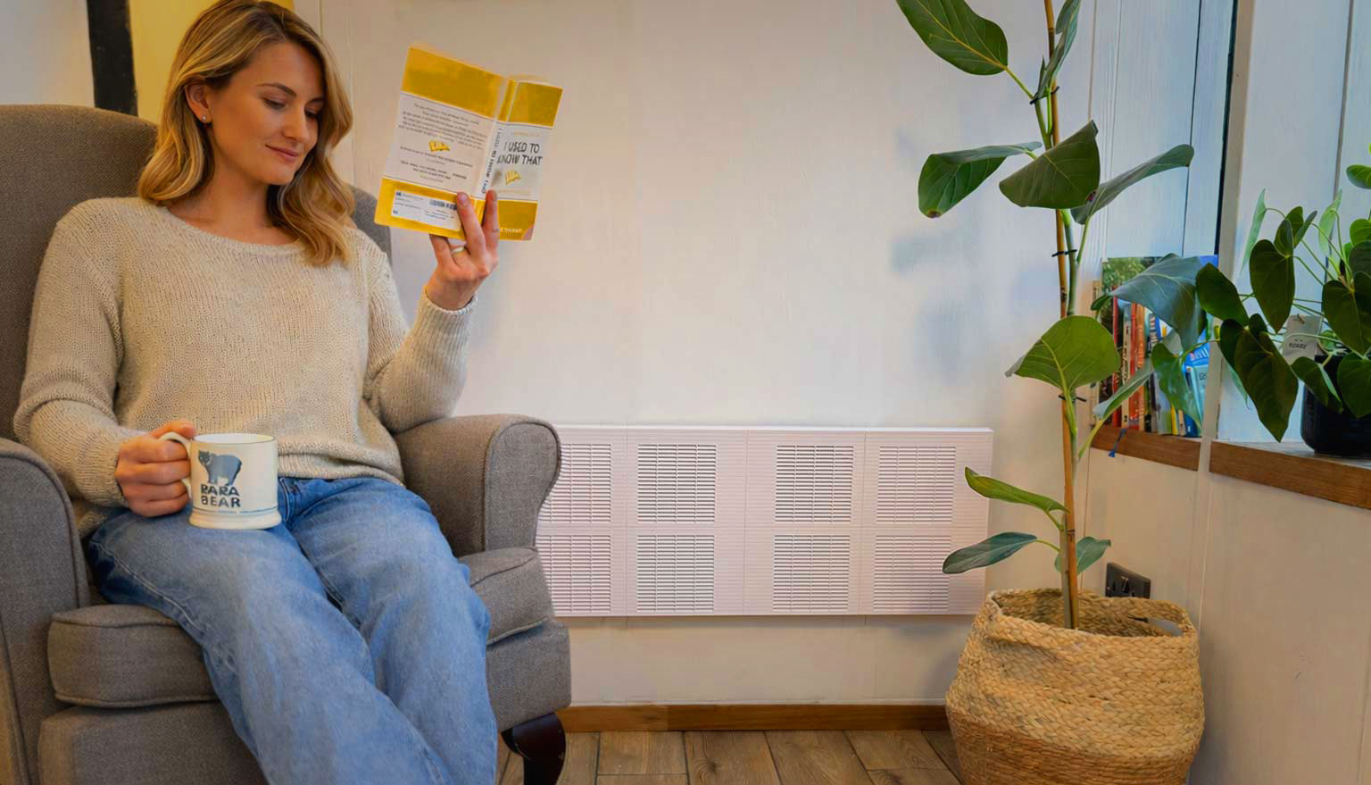 Woman sitting in a gray armchair reading a book and holding a mug that says 'PAPA BEAR' in a cozy room with wooden floor and a potted plant. Trimley in the background