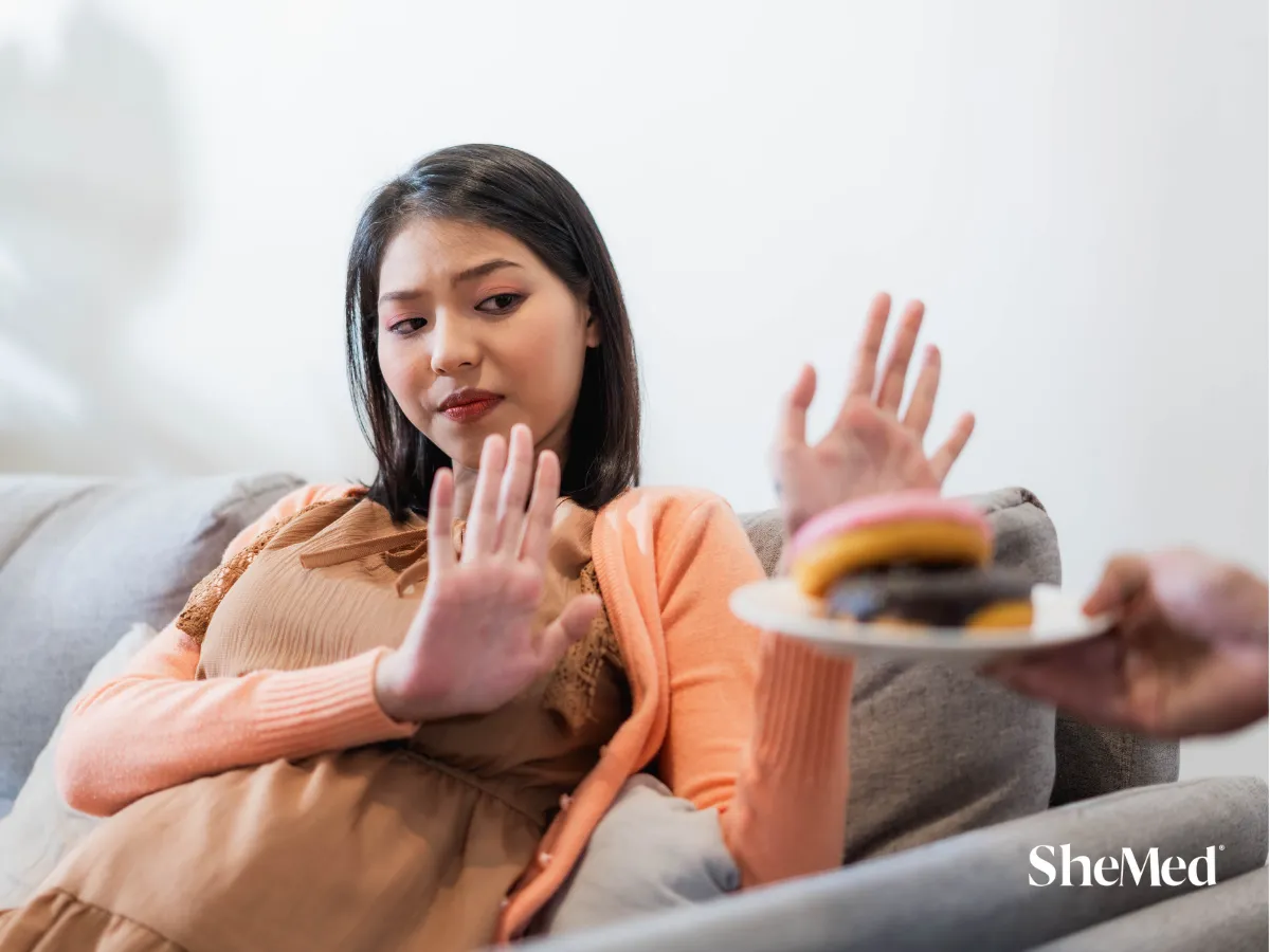 Girl sitting on a couch, politely refusing a doughnut offered by someone else, with a gentle but firm expression, with SheMed logo at the corner.