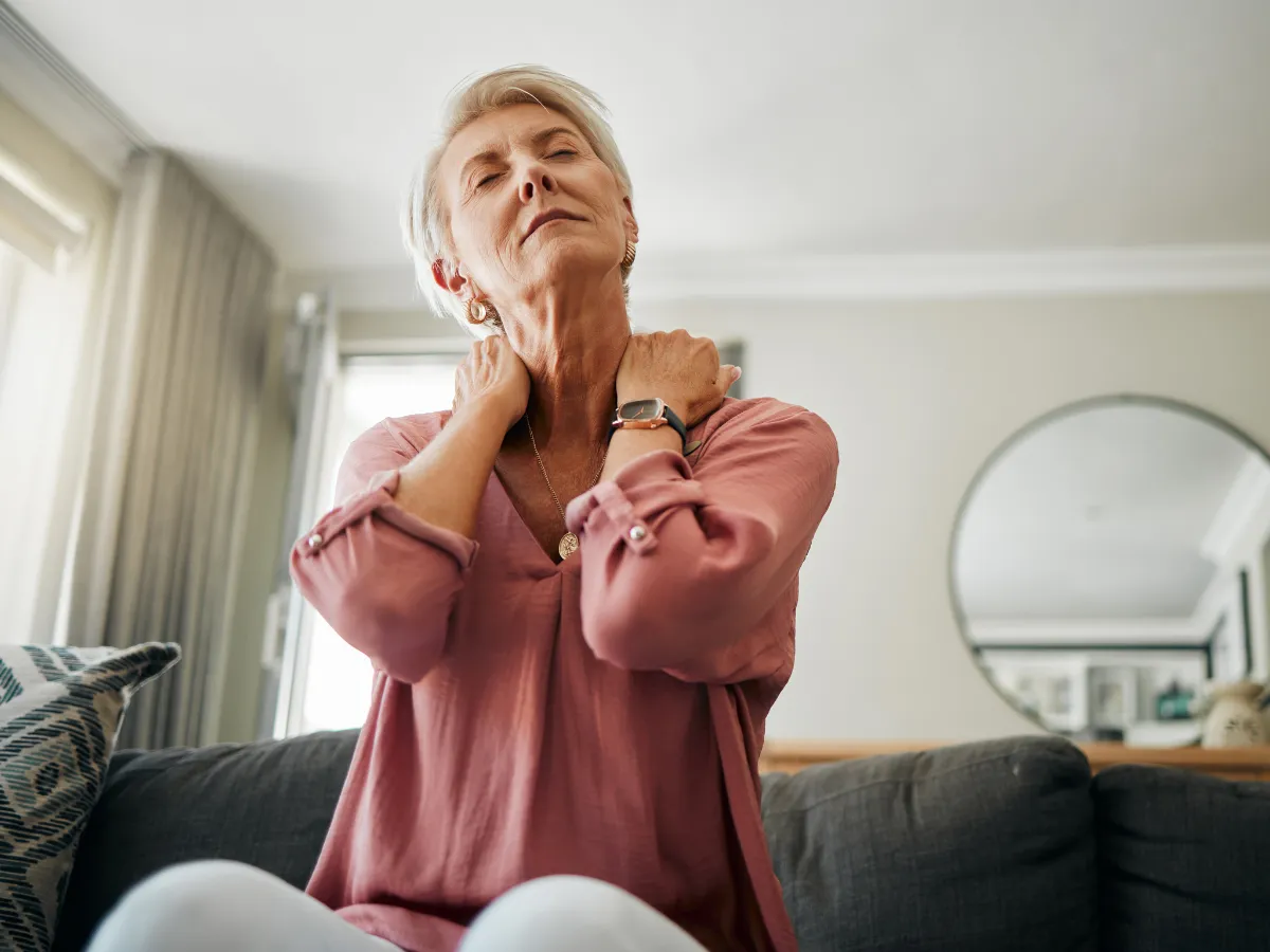 Elderly woman in a pink blouse sits on a couch, massaging her neck with a pained expression.