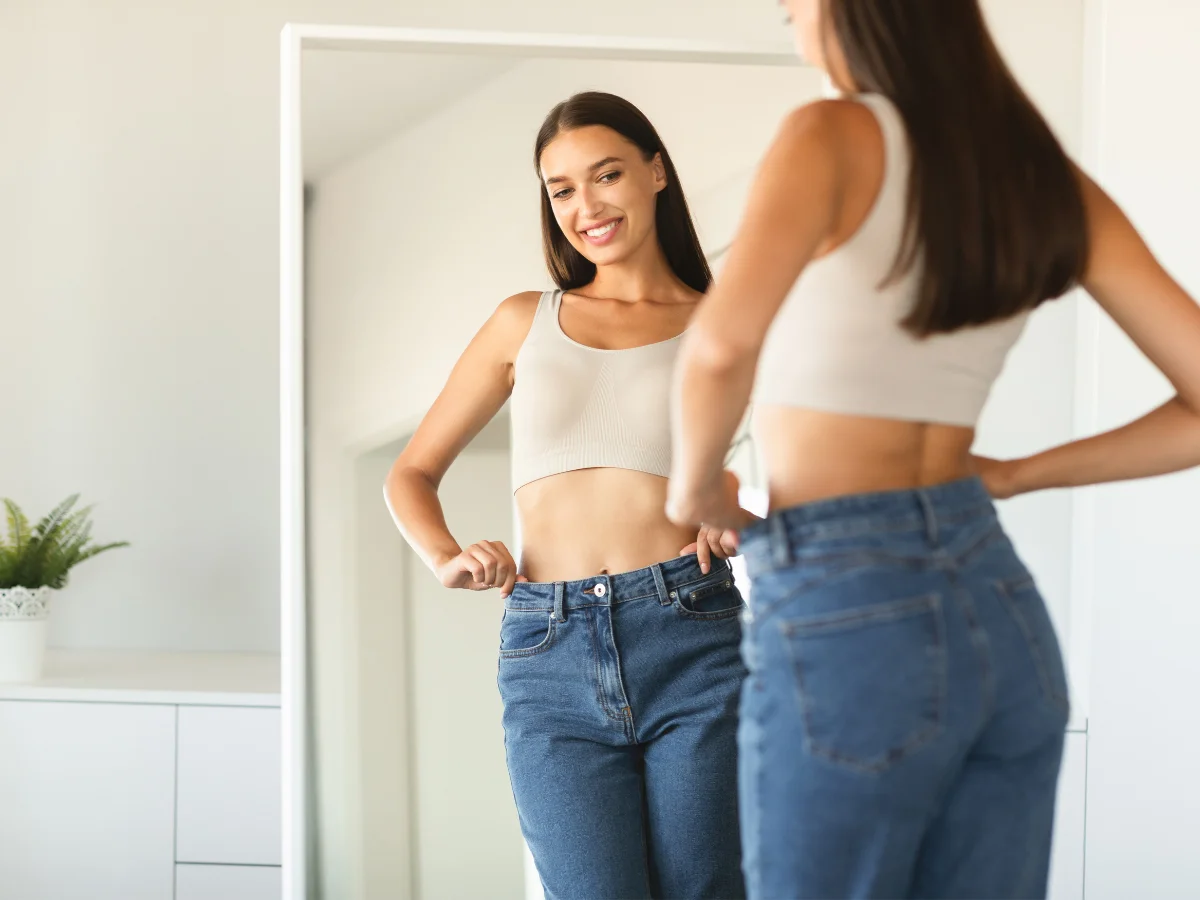  Smiling woman checking loose-fitting jeans in a mirror, reflecting healthy weight loss progress at home.