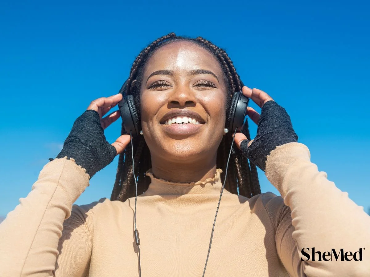 Smiling woman with box braids wearing headphones outdoors against a bright blue sky, enjoying music.