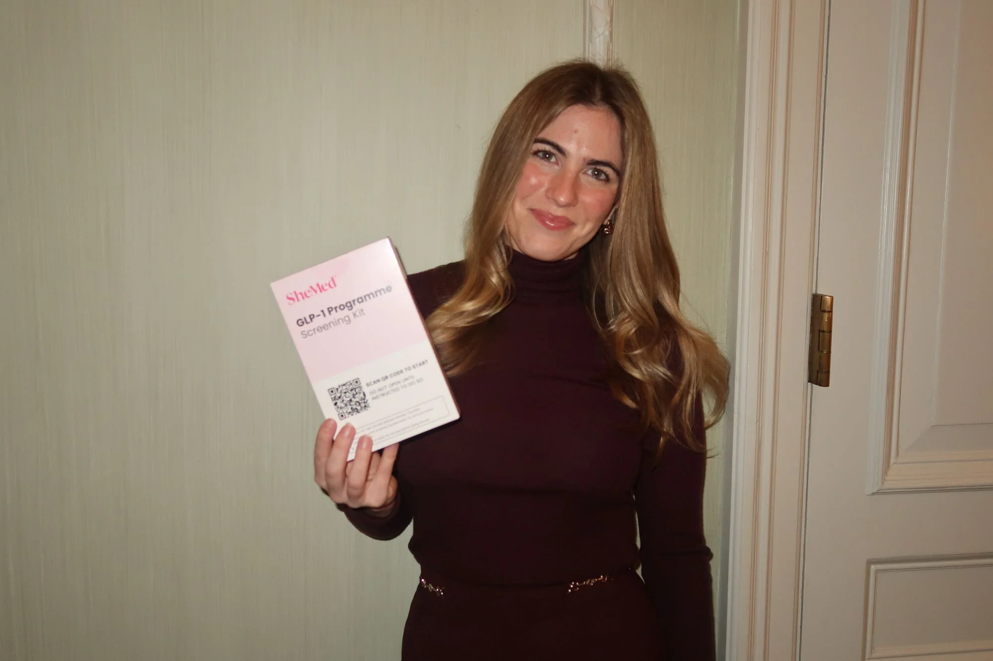 Woman in a burgundy turtleneck holding a SheMed GLP-1 Programme Screening Kit with a QR code.