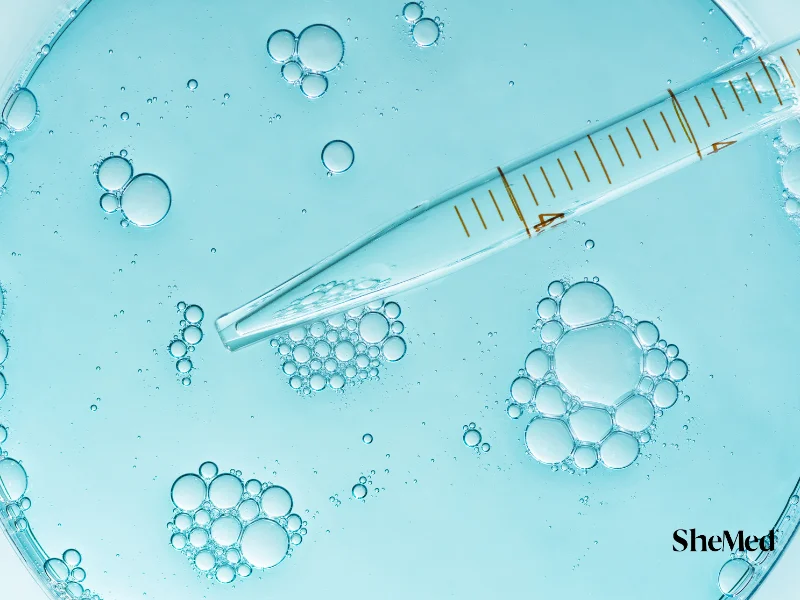 Close-up of a glass pipette over a petri dish with bubble clusters on a light blue background.