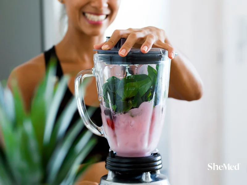 Smiling woman blending a pink berry and spinach smoothie in a glass blender for a healthy nutritious drink.