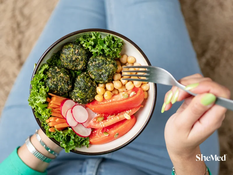 Woman holding a vegan buddha bowl with falafel, chickpeas, tomatoes, carrots, and radishes, eating with a fork.