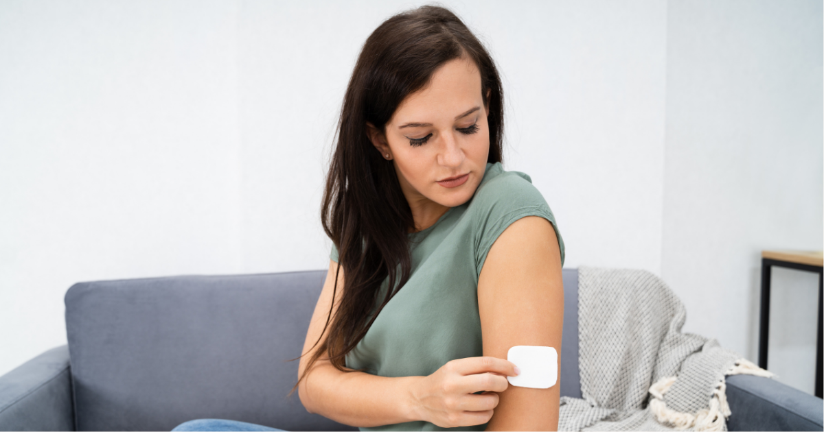 Woman seated on a sofa applying a white adhesive patch to her upper arm, representing the use of a GLP-1 patch for weight loss