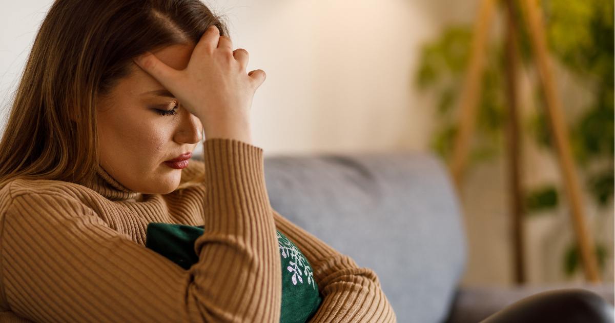 Woman sitting on sofa holding her head, experiencing headache as a side effect of GLP-1 weight loss medication
