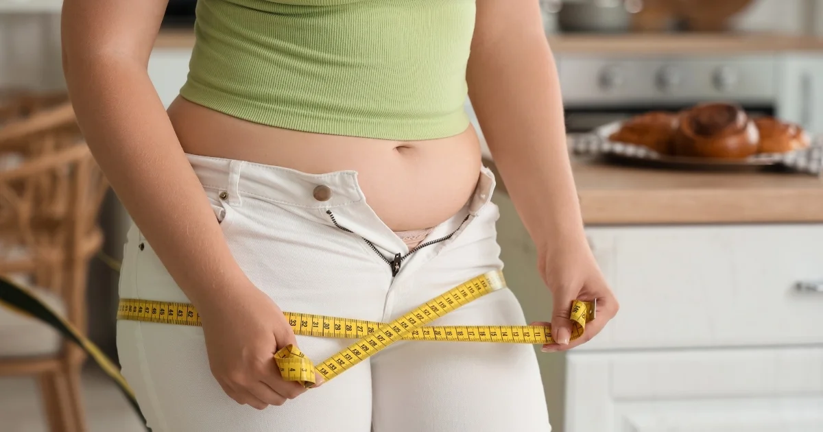 Waist measurement with tape measure, unable to zip up jeans, pastries in background, illustrating weight gain linked to PCOS and menopause.