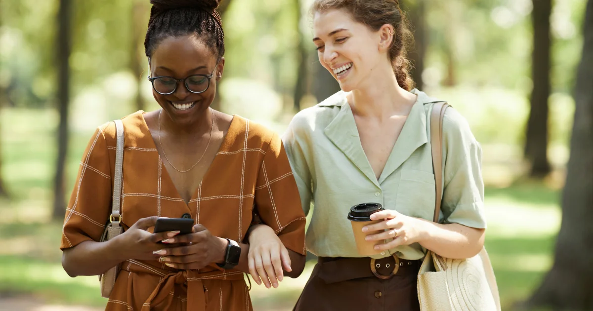 Two women walking together in a park, smiling and looking at a smartphone, representing women researching GLP-1 medication switching options between Mounjaro and Wegovy in the UK