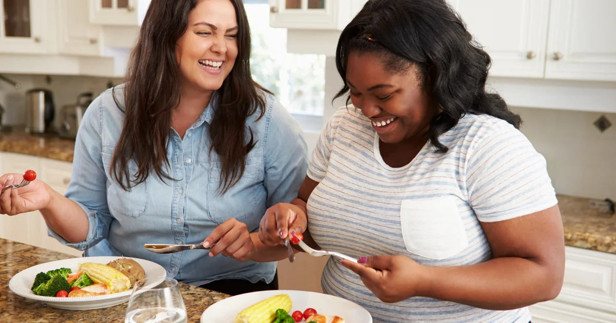 Two smiling women eating a meal of vegetables, chicken and corn together in a kitchen