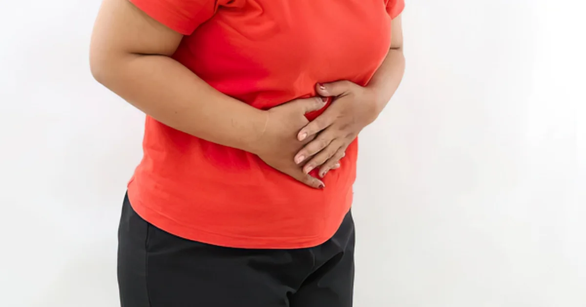 Woman in a red t-shirt and black trousers holding her stomach with both hands, showing abdominal bloating against a plain white background.