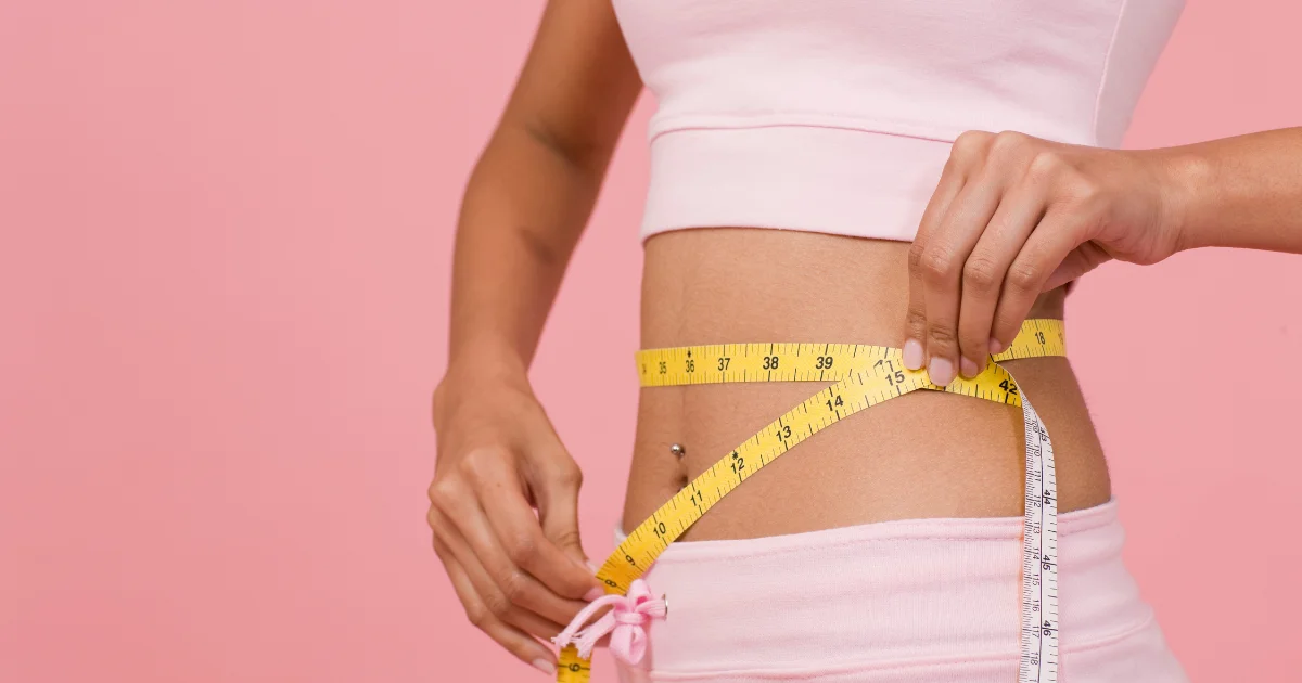 Woman in pink activewear measuring her waist with a tape measure against a pink background.