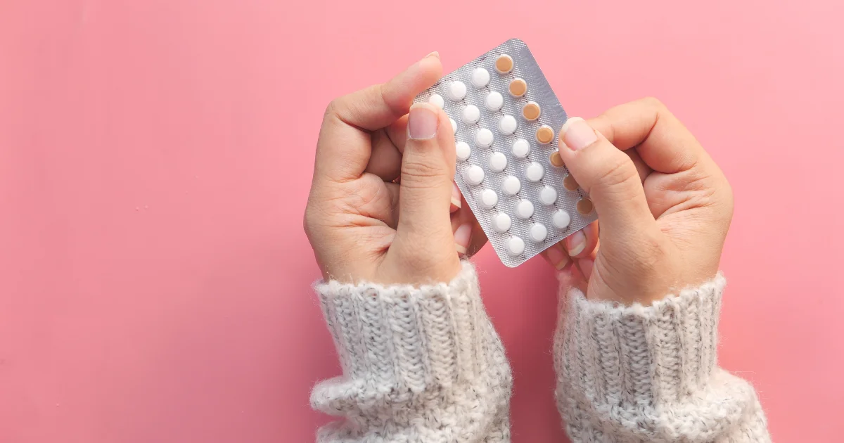 Close-up of two hands holding a blister pack of contraceptive pills against a pink background.