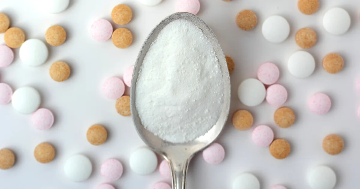 Close-up of a spoonful of white powder held above scattered white, orange and pink supplement tablets on a white background.
