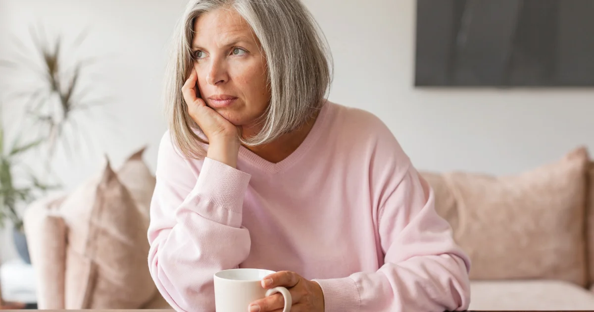 Woman with grey hair resting her chin on her hand, holding a mug and looking thoughtfully into the distance while seated indoors.