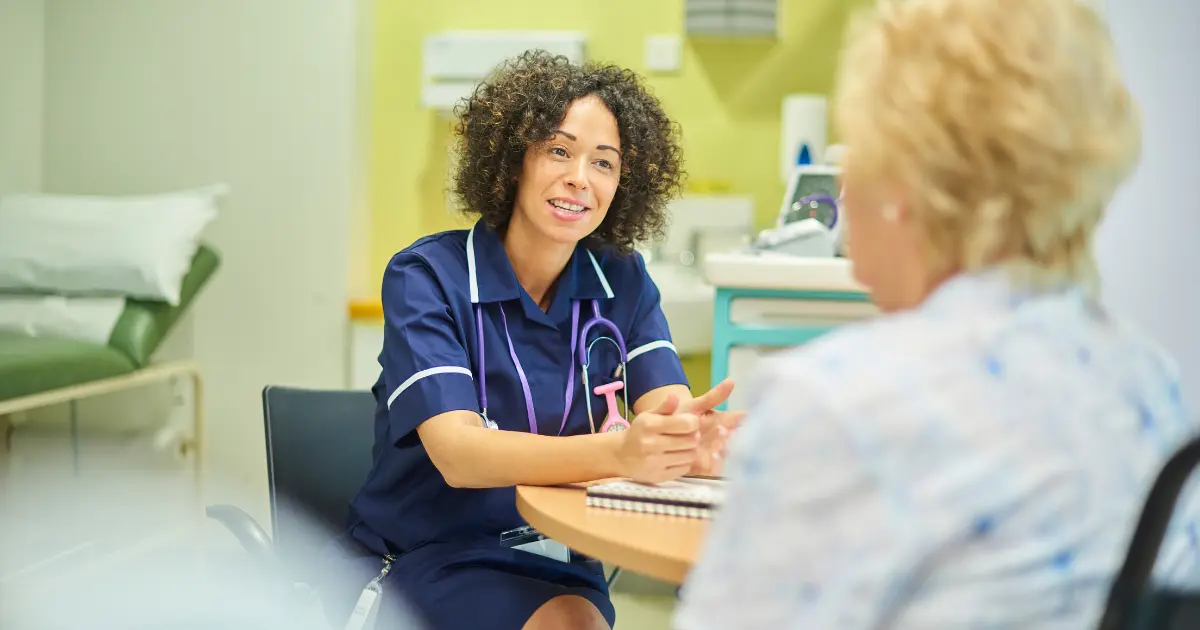 A nurse in navy scrubs with a stethoscope talking with a patient at a table in a clinical consultation room.
