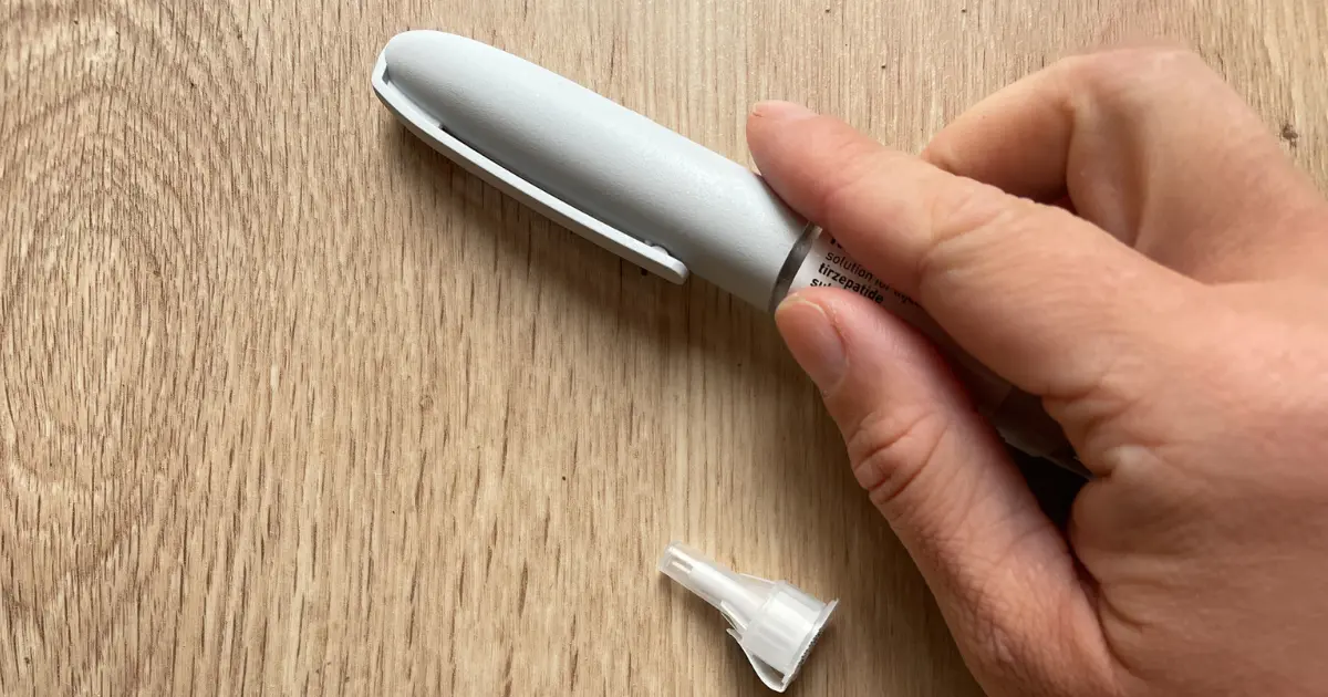 Close-up of a hand holding a Mounjaro injection pen with the label reading “tirzepatide” visible, with the pen cap removed on a wooden surface.