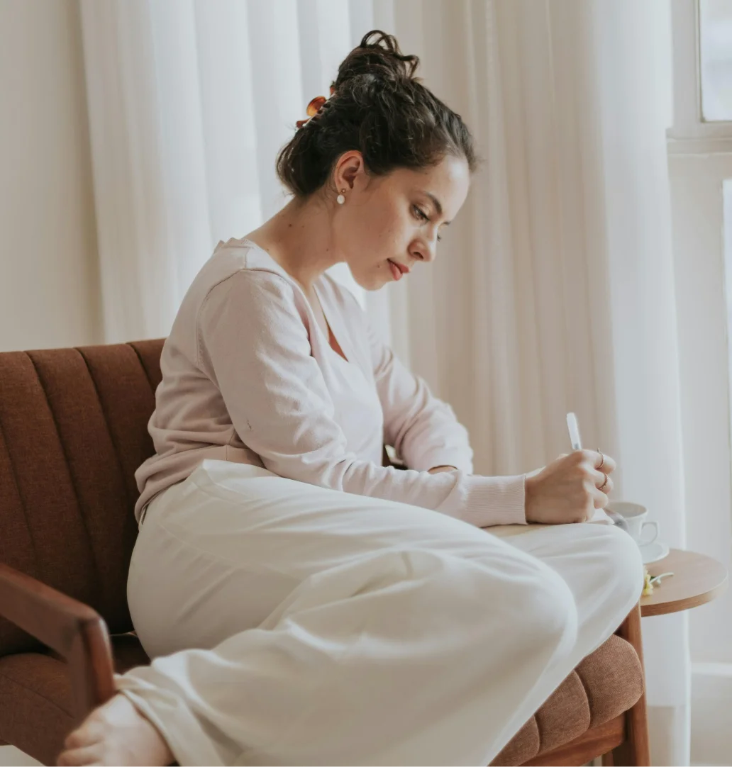 Young woman with hair tied back sitting on a brown chair by white curtains, writing with a pen on paper.