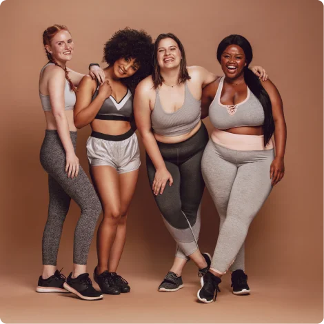 Four diverse women in athletic wear smiling and standing closely together against a brown background.