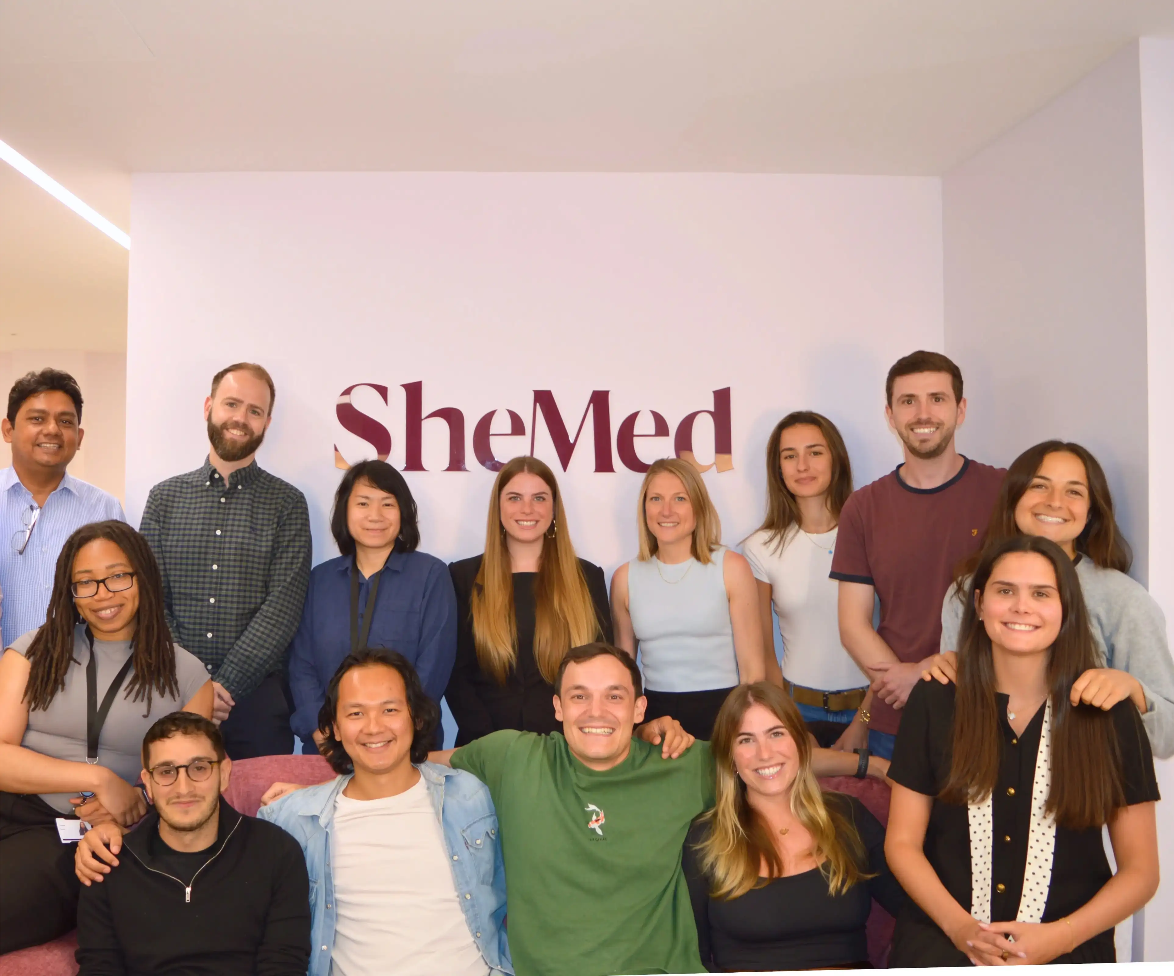 Group photo of a diverse SheMed team smiling in front of a wall with the SheMed logo.