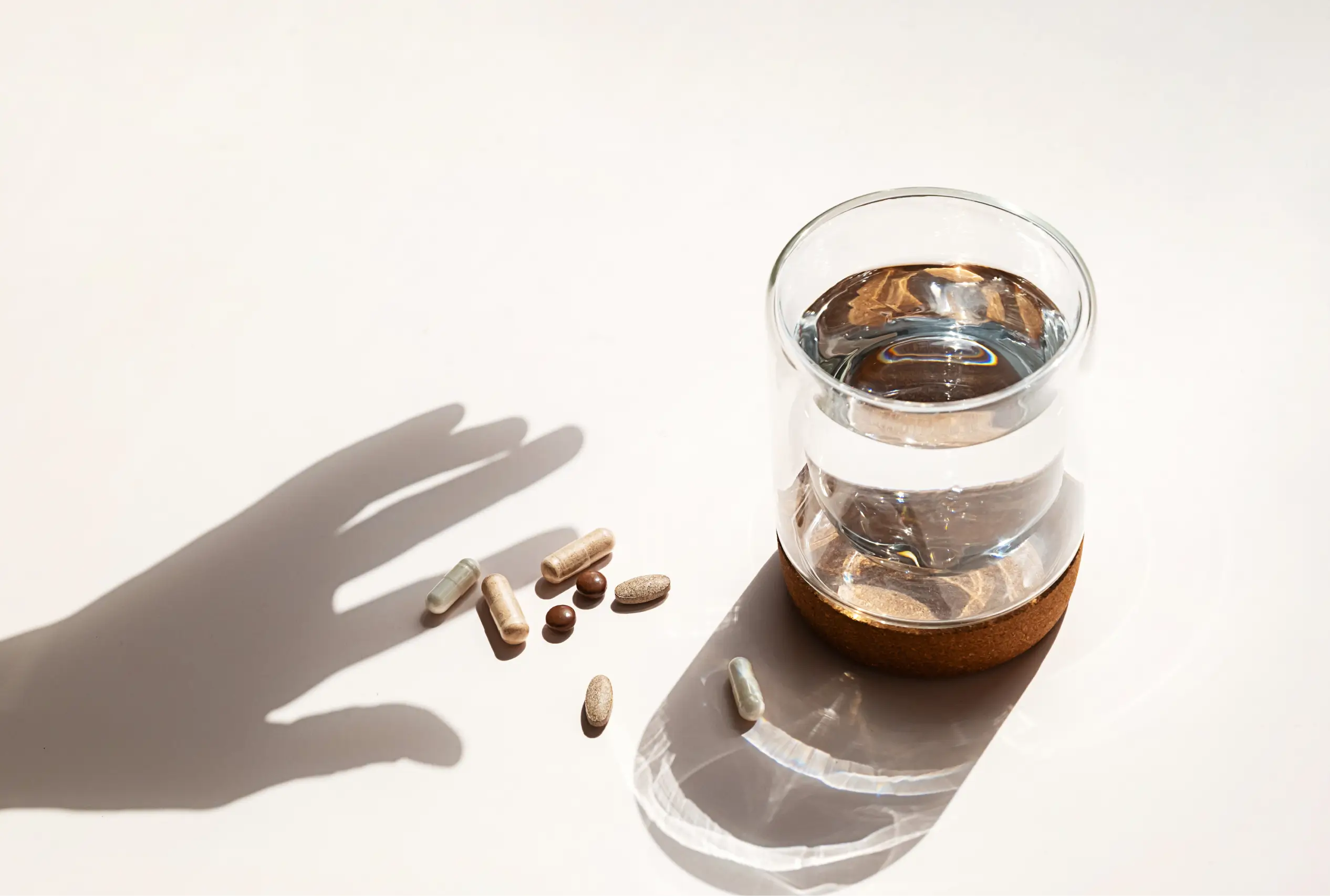 Shadow of a hand reaching toward several capsules and tablets next to a glass of water on a white surface.